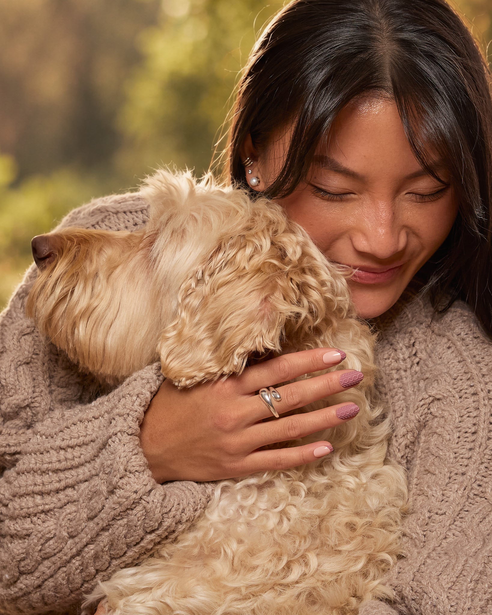 A woman in a cozy taupe knit sweater holds a fluffy cream-colored dog close to her face. She is wearing a silver ring with a clear central gemstone on her finger.