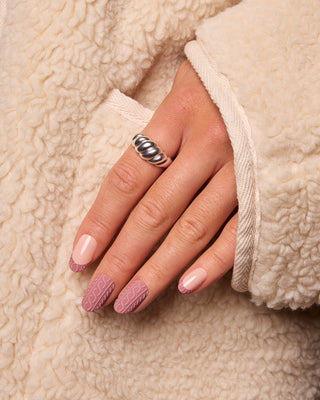 A close-up of a hand wearing a textured silver ring, resting on a cream-colored sherpa jacket. The nails are manicured with a mix of solid pale pink and pink textured nail art.
