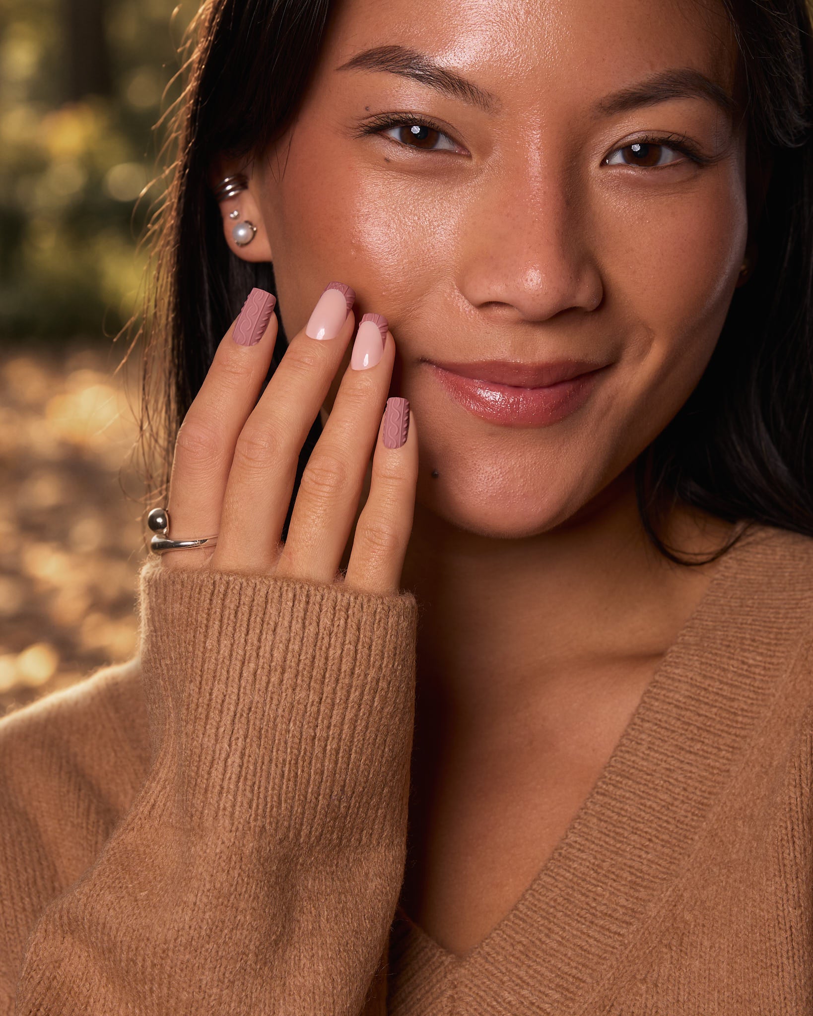 Close-up portrait of a woman outdoors wearing a tan knit sweater, with her hand near her face. She has pinkish nude nail polish, a silver ring, and a pearl earring.