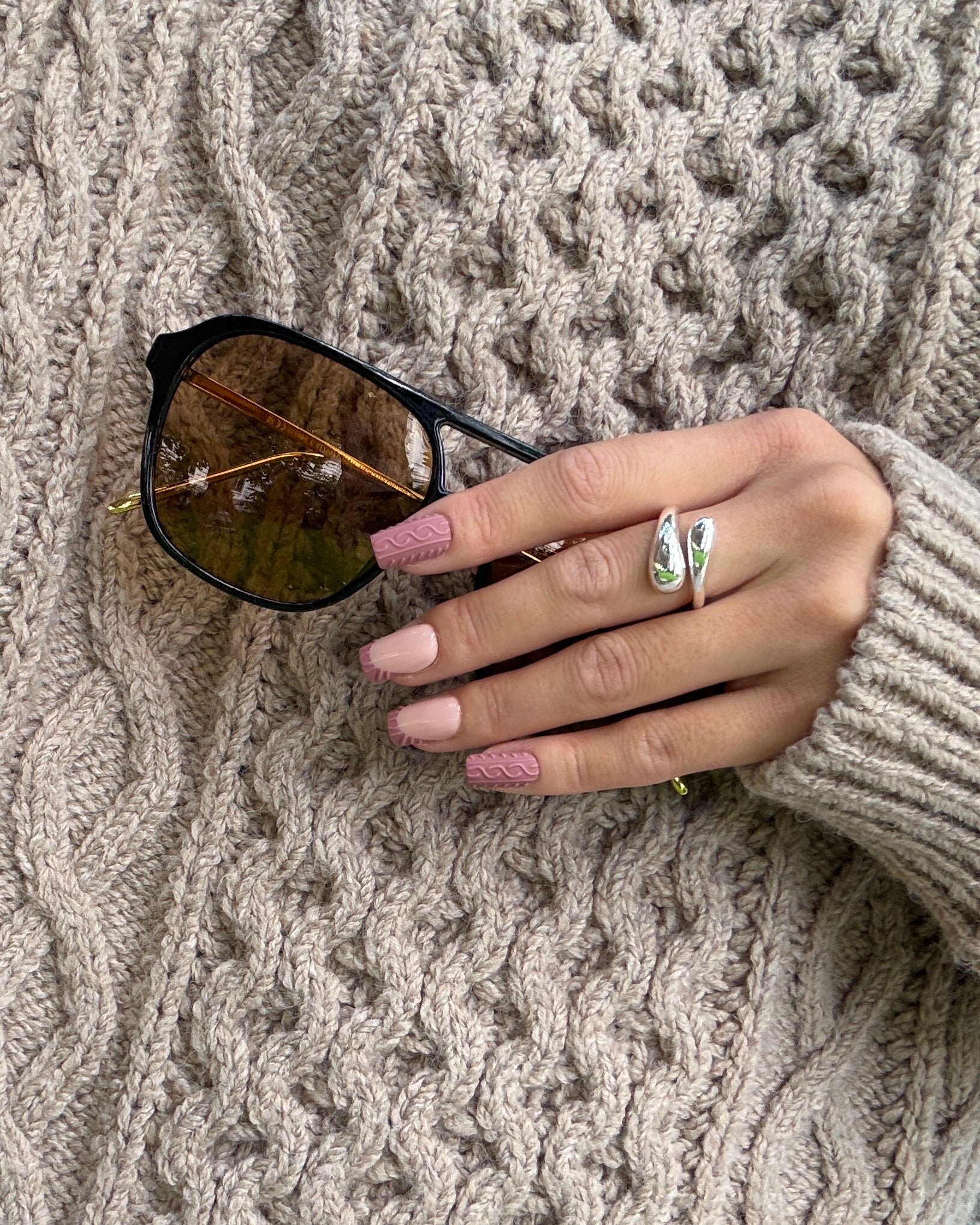A close-up of a hand resting on a chunky beige cable-knit sweater, wearing a silver split-ring ring with green and white stones. The hand holds a pair of black-framed sunglasses with gold-toned lenses.