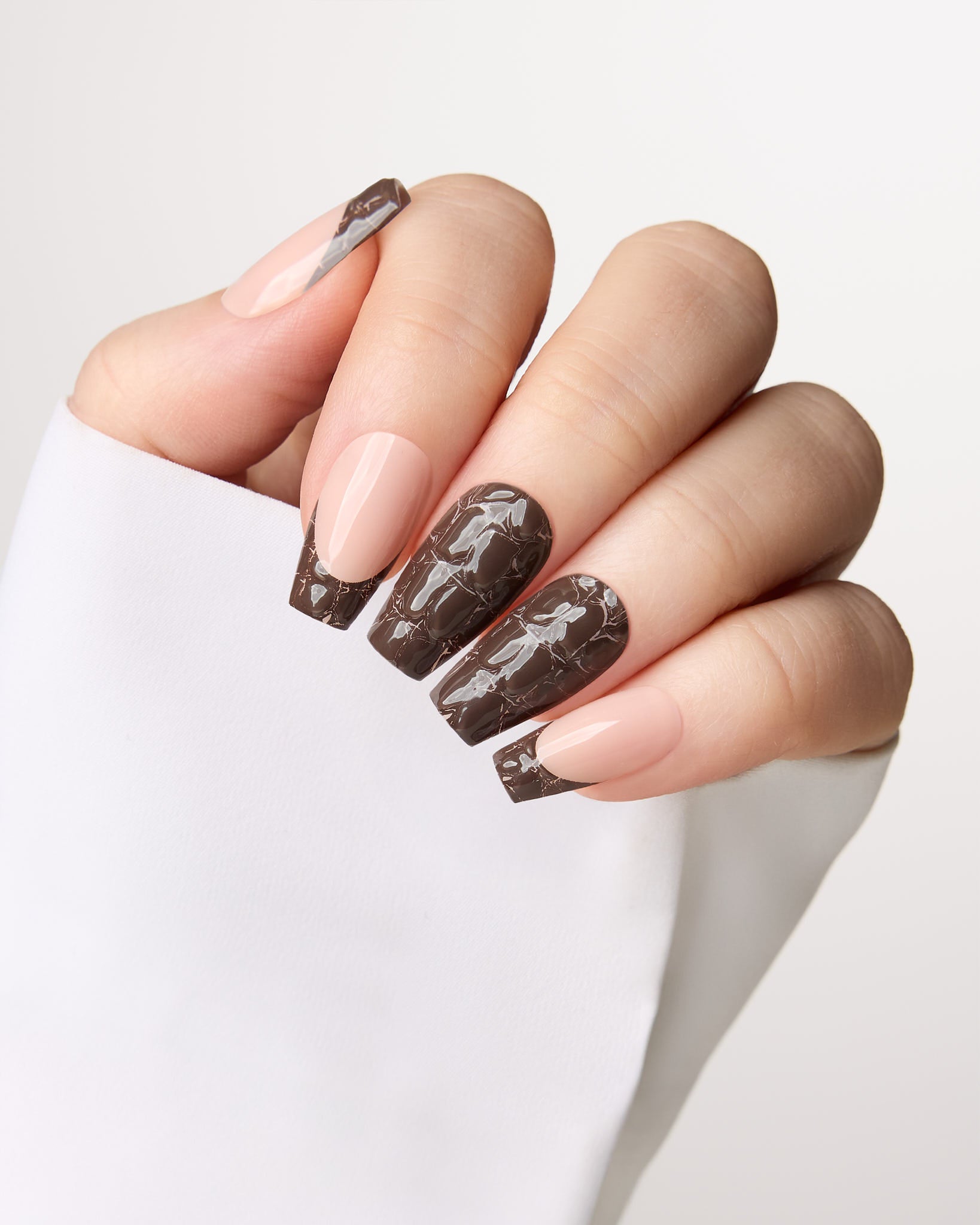 A close-up of a hand with long almond-shaped nails styled in a glossy dark brown and nude pink French manicure, wearing a white sleeve against a light gray background.
