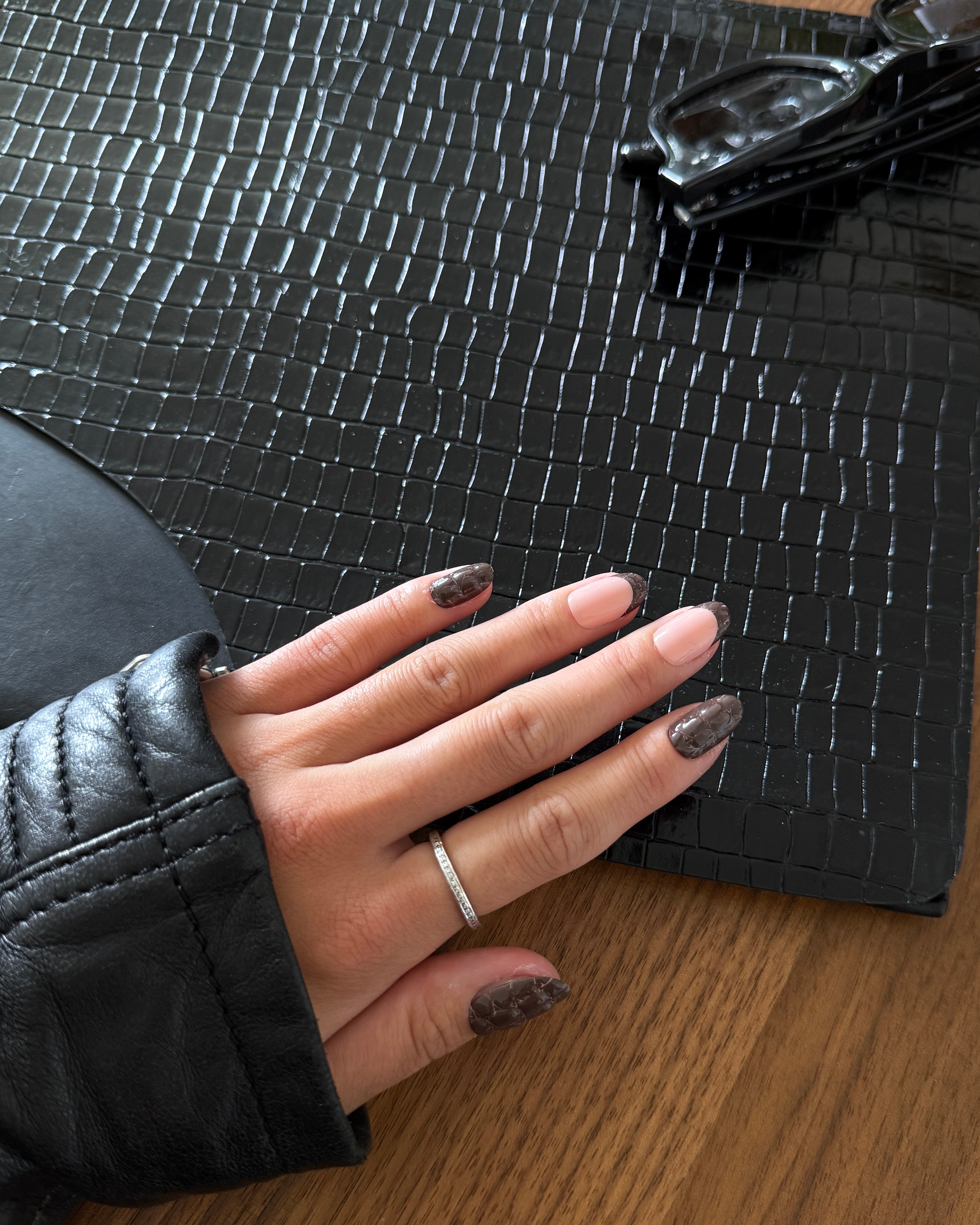 A close-up of a hand with dark brown glittery nail polish resting on a black crocodile-embossed leather surface, wearing a thin silver band ring. The hand is also wearing a black leather sleeve, and a wooden table is visible underneath.