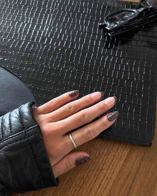 A close-up of a hand with dark brown glittery nail polish resting on a black crocodile-embossed leather surface, wearing a thin silver band ring. The hand is also wearing a black leather sleeve, and a wooden table is visible underneath.