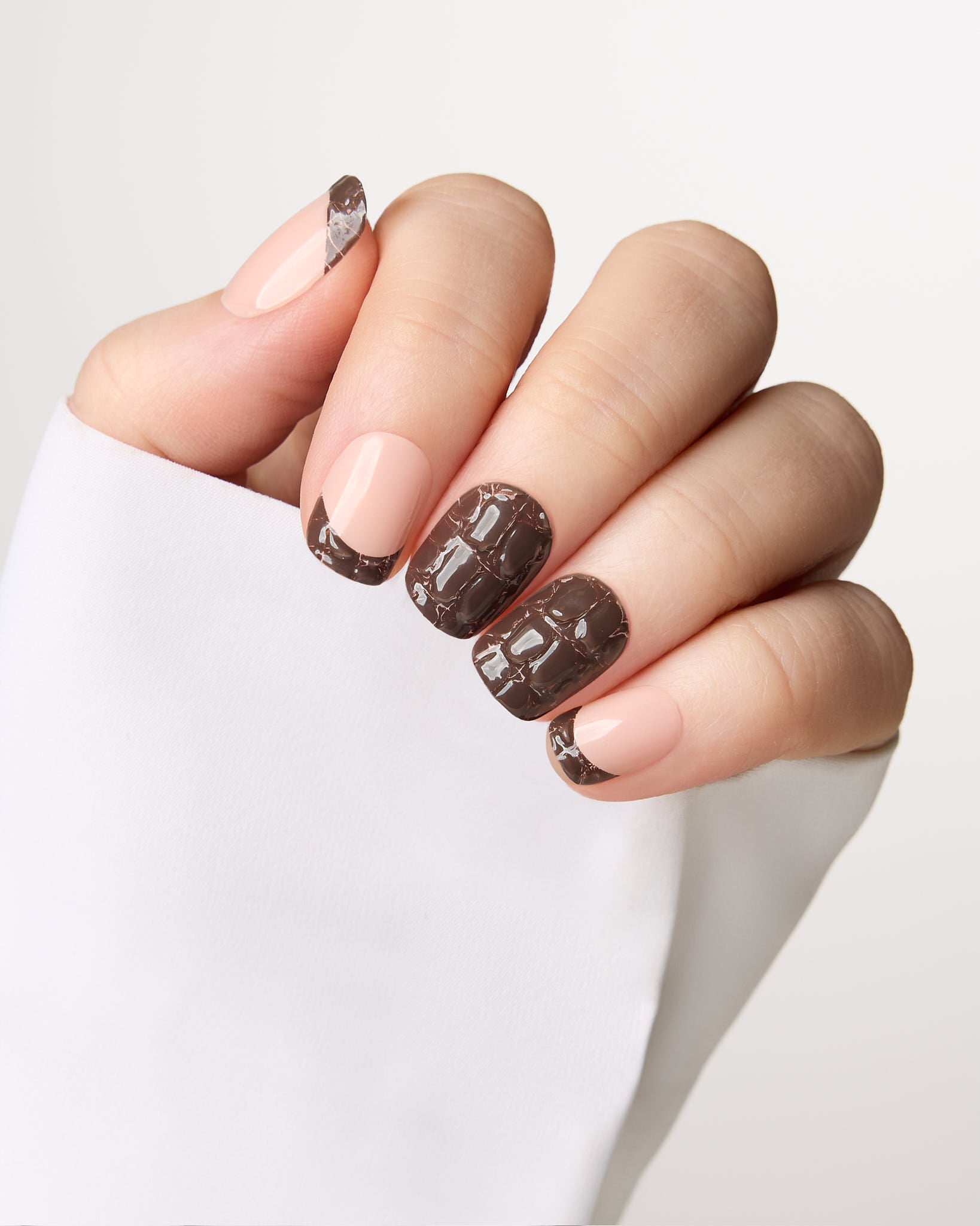 A close-up of a hand with manicured nails, featuring three dark brown glossy crocodile-embossed patterned nails and three nude pink nails, resting on a white sleeve against a light gray background.