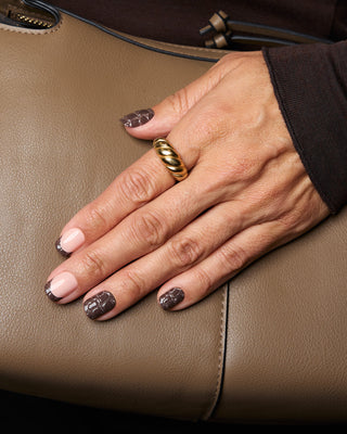 A close-up of a hand resting on a brown textured leather surface, wearing a chunky twisted gold ring on the ring finger. The nails are styled with alternating light pink and dark brown glittery manicures.