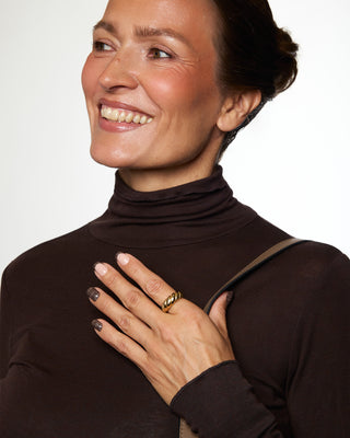 A close-up of a woman wearing a dark brown ribbed turtleneck sweater and a gold ring, smiling and looking to the side against a light background.