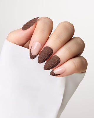 A close-up of a hand with manicured nails styled in a dark brown chocolate pattern on the outer sides and a pale nude pink polish on the inner sides, wearing a white sleeve.