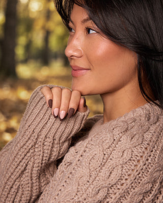 A close-up lifestyle shot of a woman in profile wearing a chunky, cable-knit brown sweater, set against a soft-focus autumn forest background.