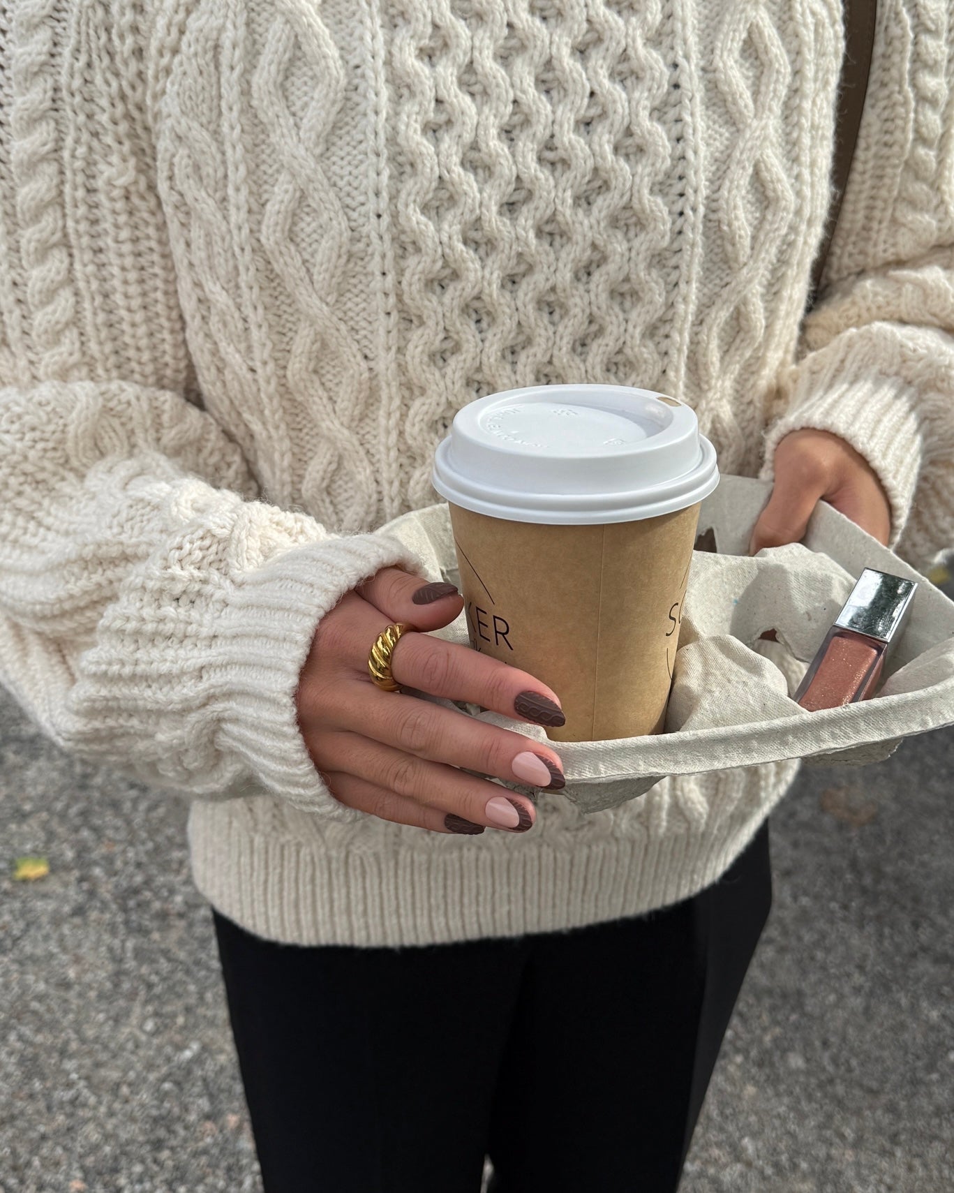 A person wearing a chunky cream cable-knit sweater holds a paper coffee cup in a beige tote bag outdoors on a paved surface.