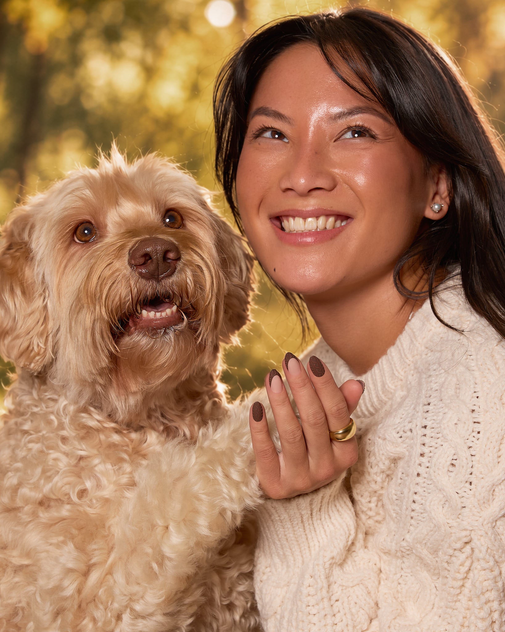 A smiling woman in a cream cable-knit sweater holds a fluffy cream-colored dog against a warm, blurred golden outdoor background.